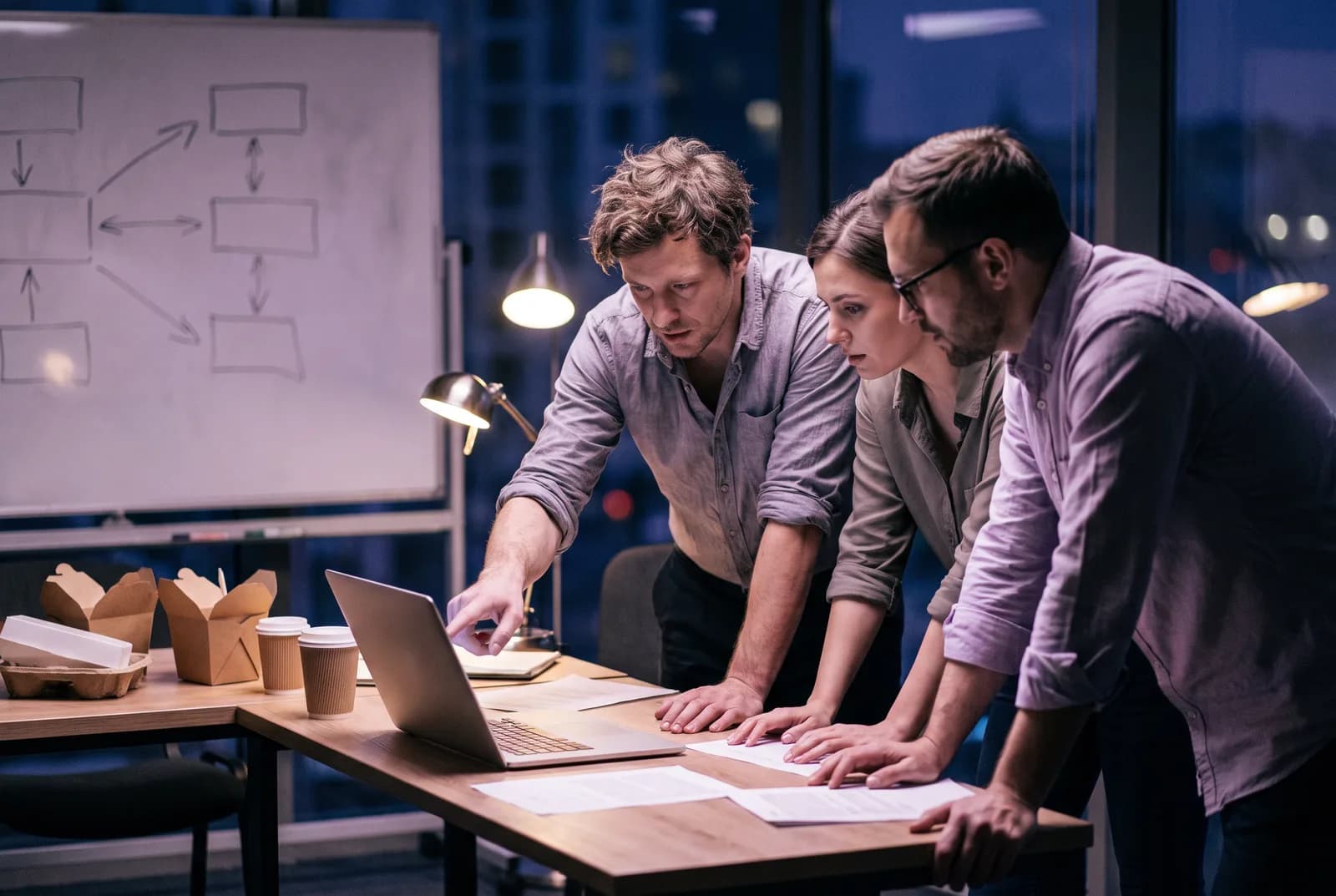 Three colleagues leaning over a laptop at night by a whiteboard, one pointing at the screen
