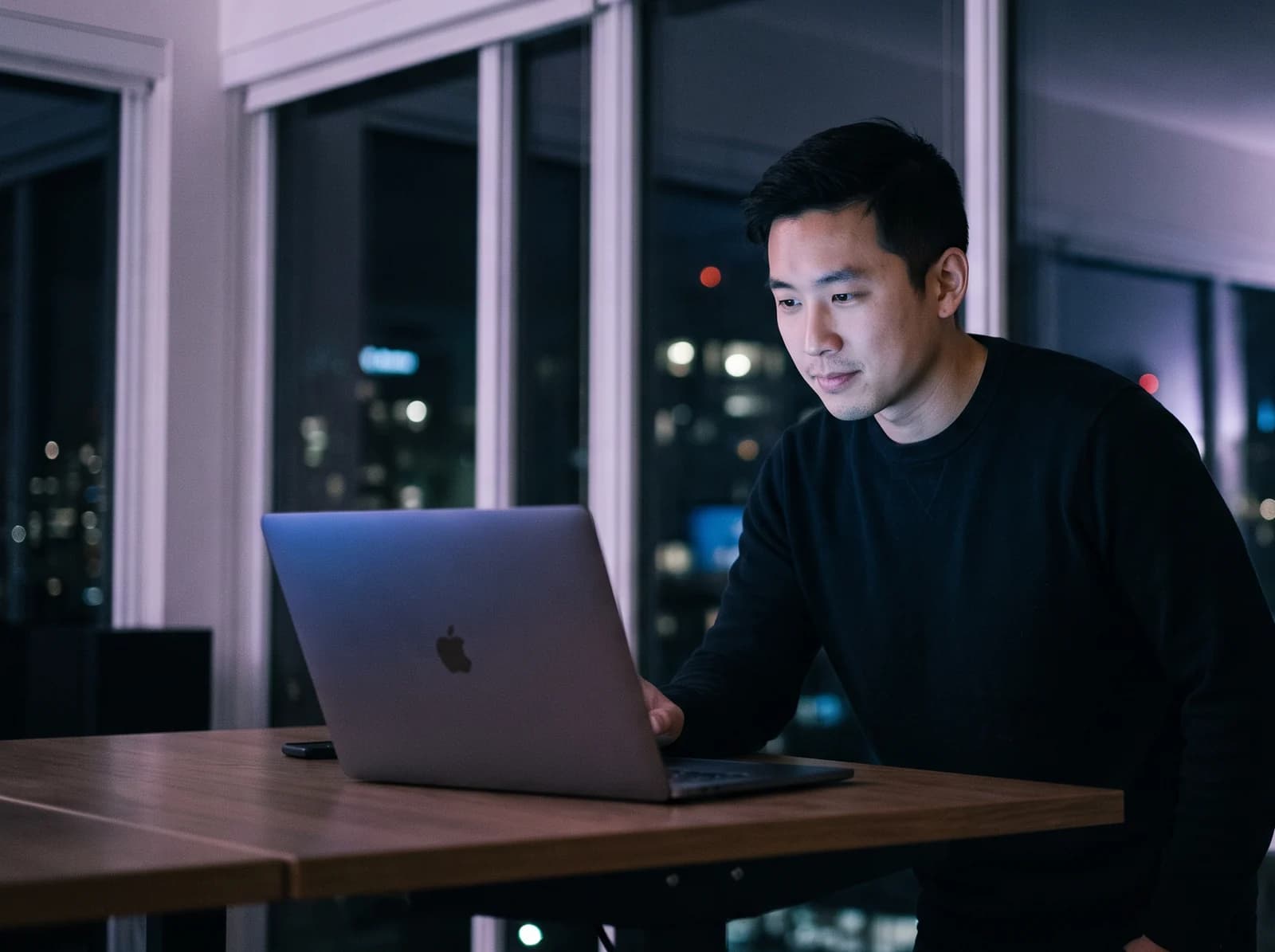 A man standing at a table by a night window over the city, working in his books on a laptop