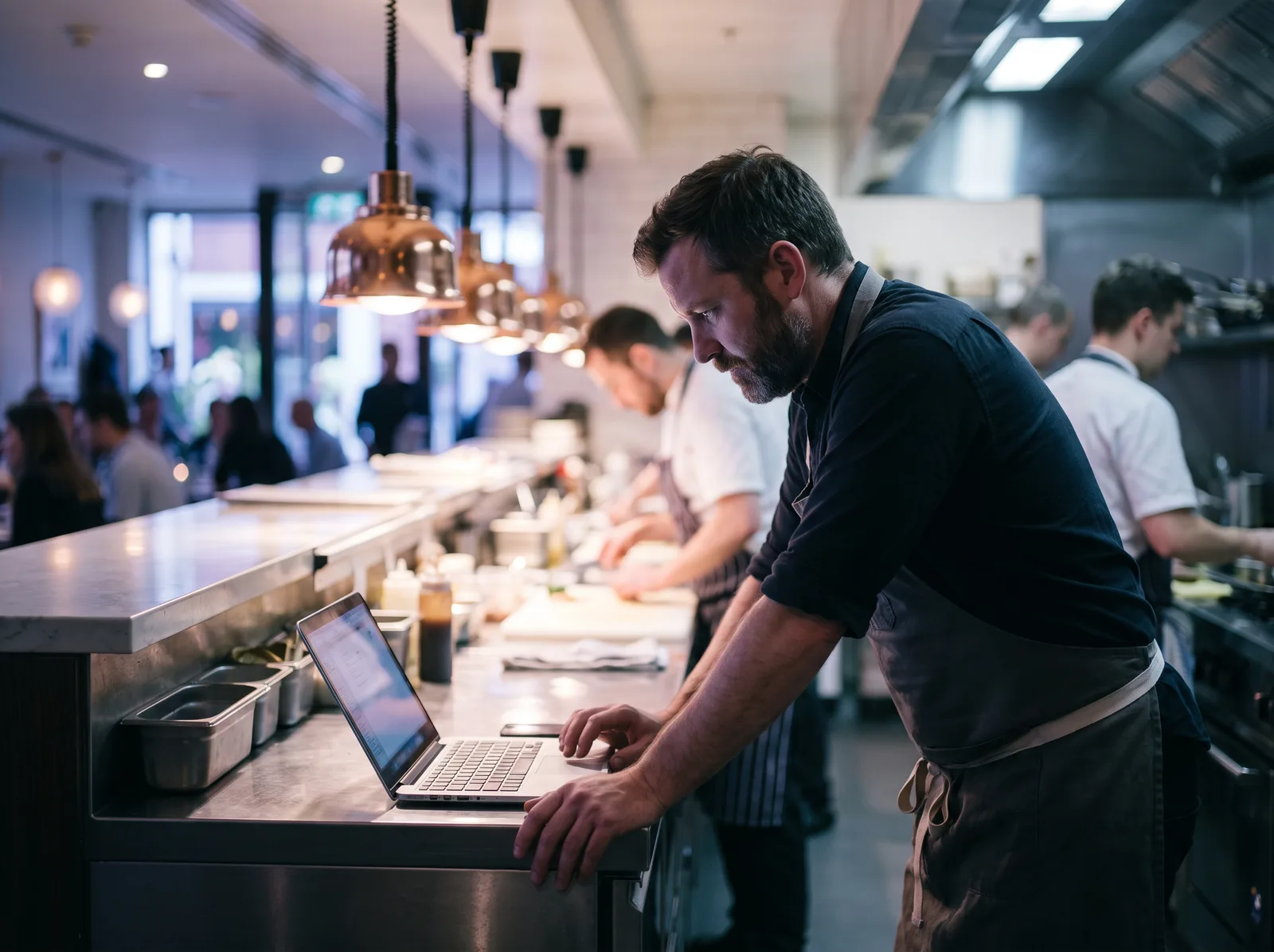 A chef at the pass of a busy restaurant kitchen, leaning over his laptop
