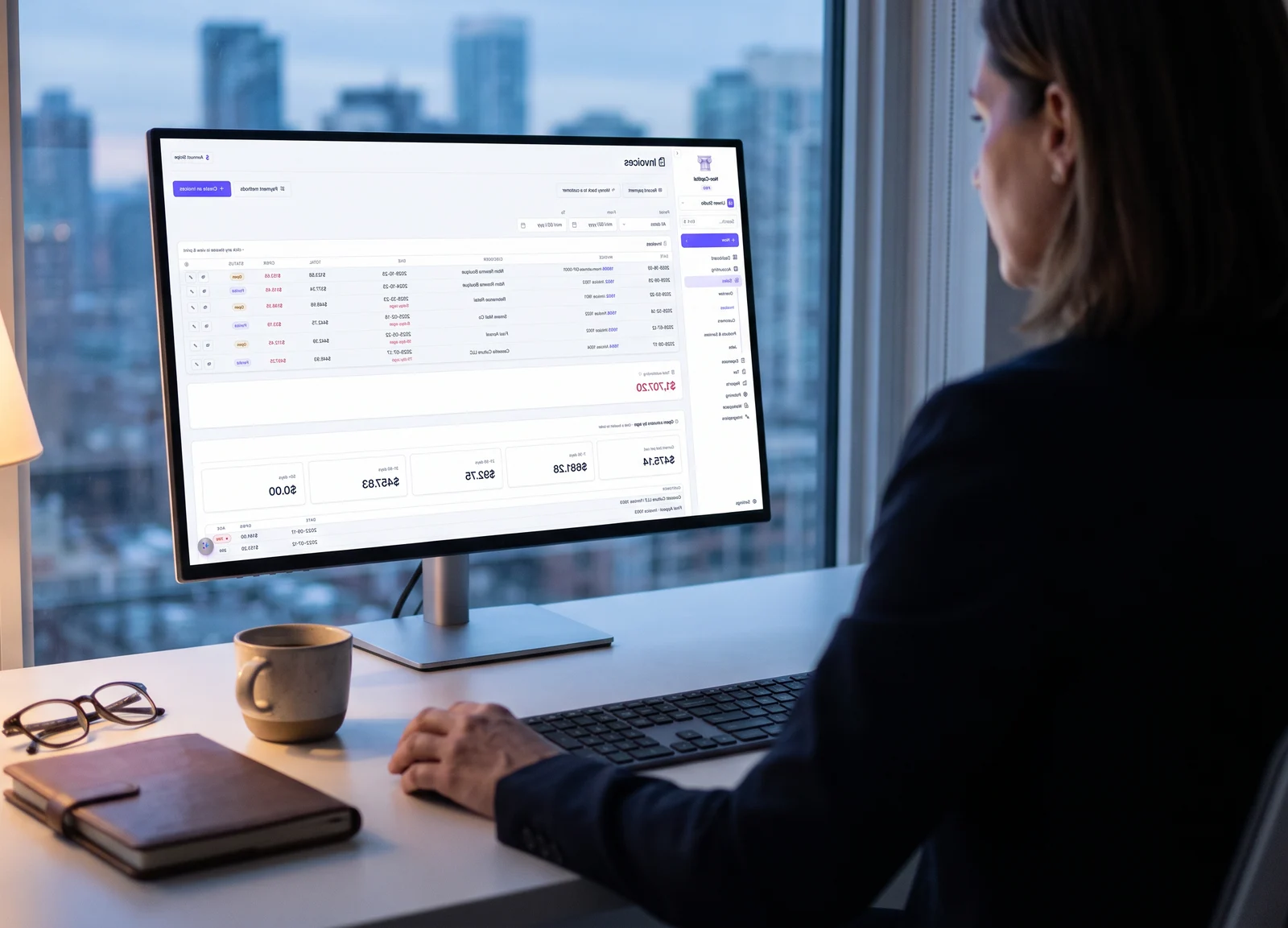 A woman at her desk by a high window at dusk, working in Neo-Capital's invoices on a large monitor