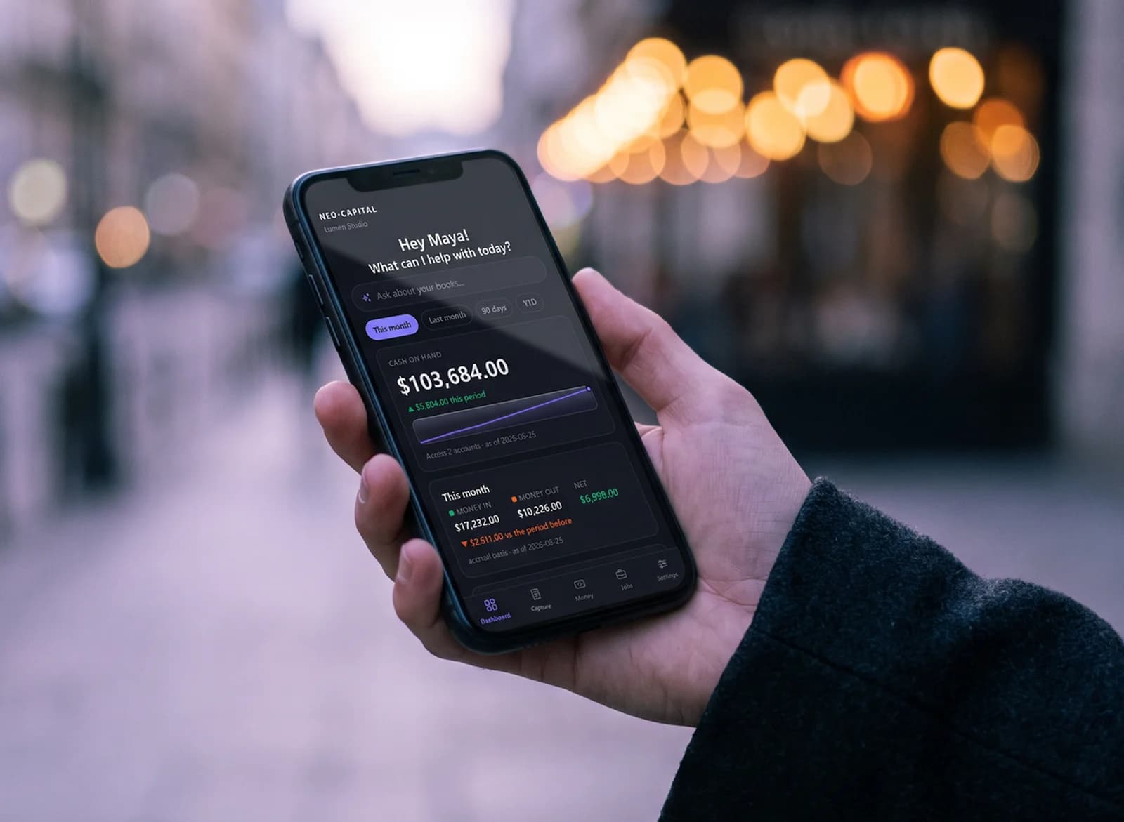 A hand holding a phone showing the Neo-Capital app's dashboard, on a city street at dusk with café lights behind